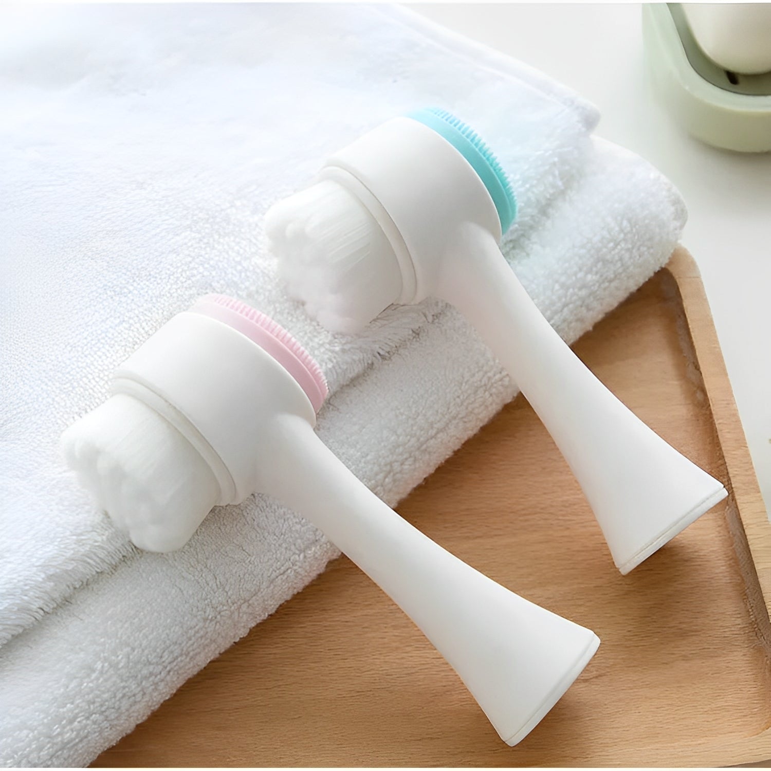 Två Elevkassan Dubbelsidig Ansiktsborste (2-pack) med långa handtag, perfekta för mild rengöring och skonsam exfoliering, vilar på en vikt vit handduk - den ena med rosa accent, den andra blå - på en träyta.