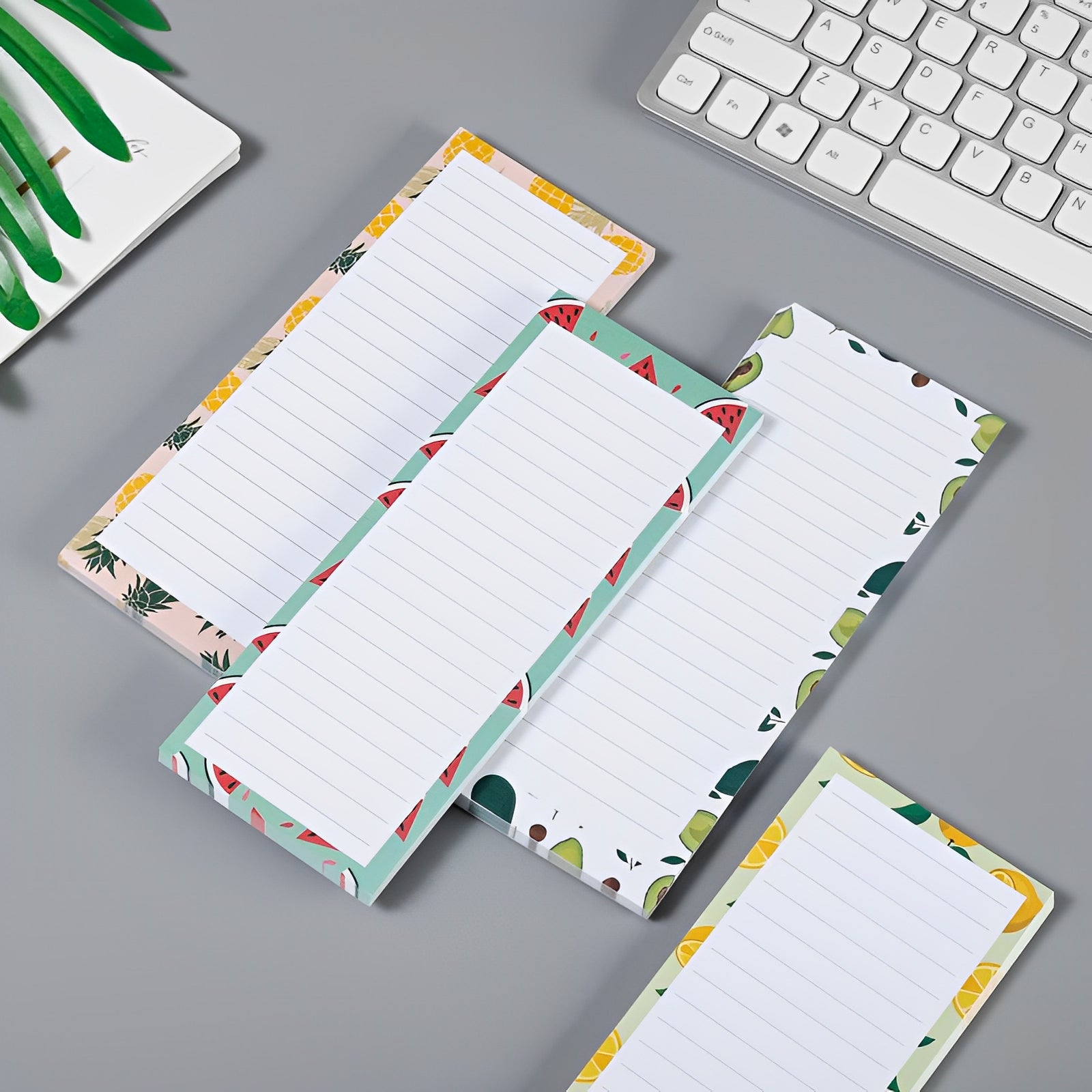 Fyra Elevkassan Magnetblock för Kylskåpet (4-pack) med färgglada fruktkanter ligger på ett skrivbord vid ett tangentbord och en växt. Varje fodrat anteckningsblock är perfekt för att anteckna inköpslista eller använda som kylskåpsmagnetblock.