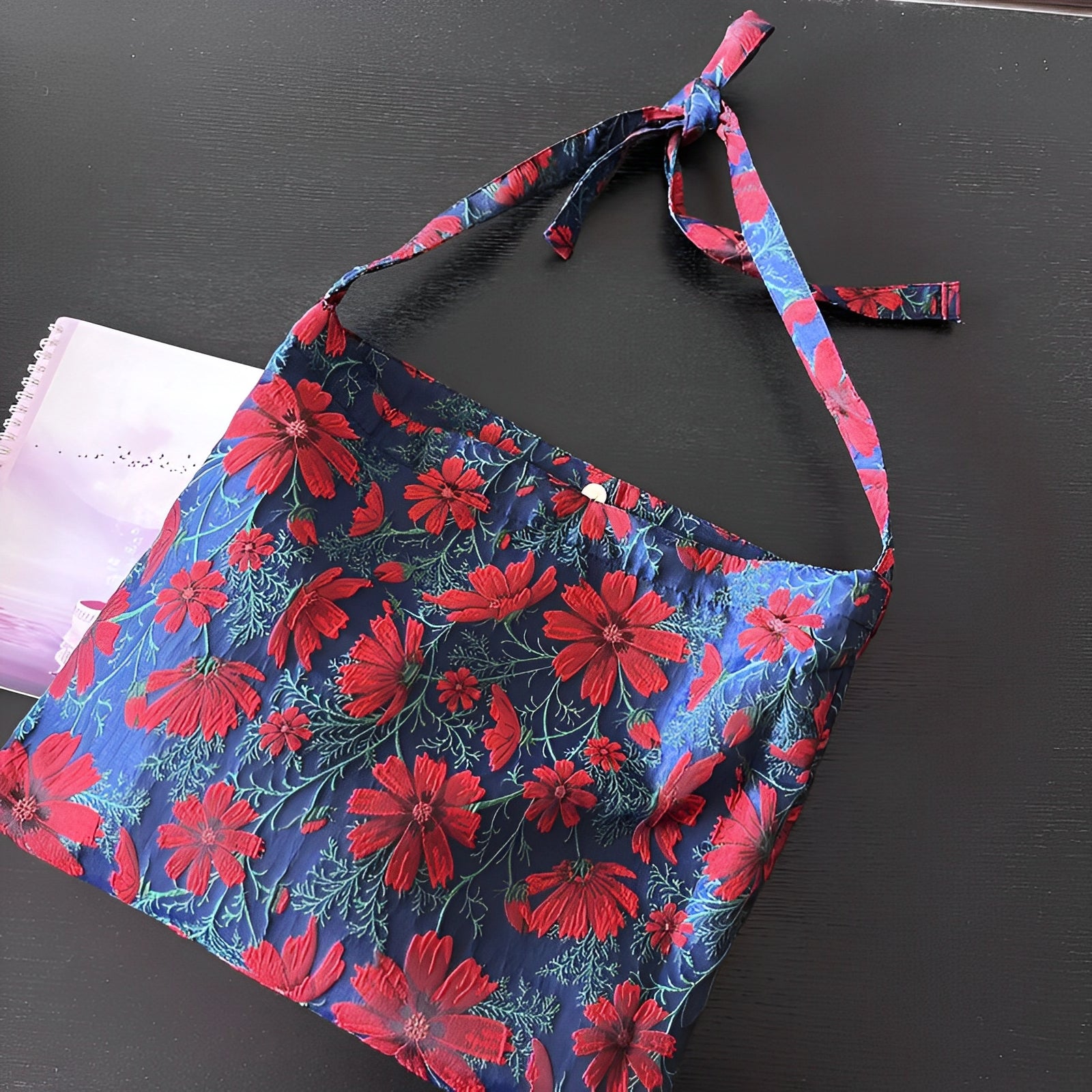 En Blommig Tygväska - 34x38cm från Elevkassan, med en blå bas med rött blommönster och ett knutet justerbart band, ligger på ett mörkt bord bredvid en delvis synlig lila spiralbok.