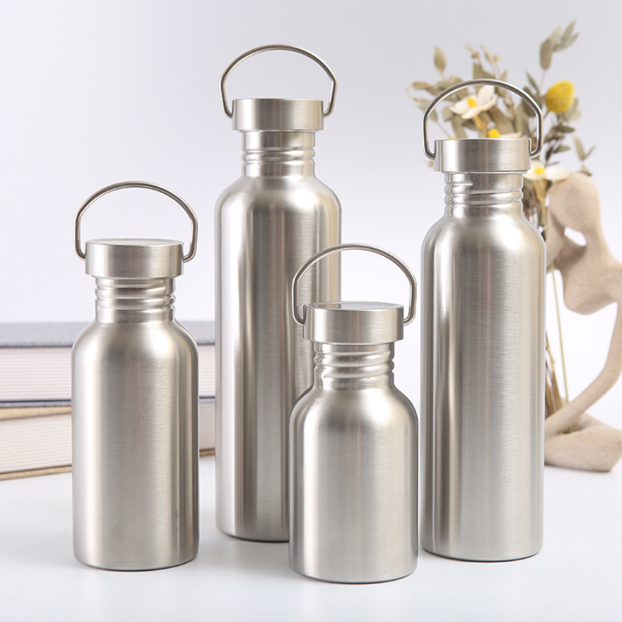 Fyra Elevkassan Metallflaska i Rostfritt Stål med stilrena handtag står på en vit yta, omgivna av staplade böcker, torkade blommor och en beige abstrakt skulptur i bakgrunden.